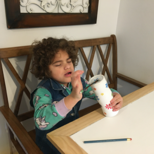 A 9-year-old girl with Angelman syndrome sits at a table in a house holding a container decorated with white paper and colorful flowers. She pushes a pencil into a hole at the top of the container to practice her fine motor skills. 